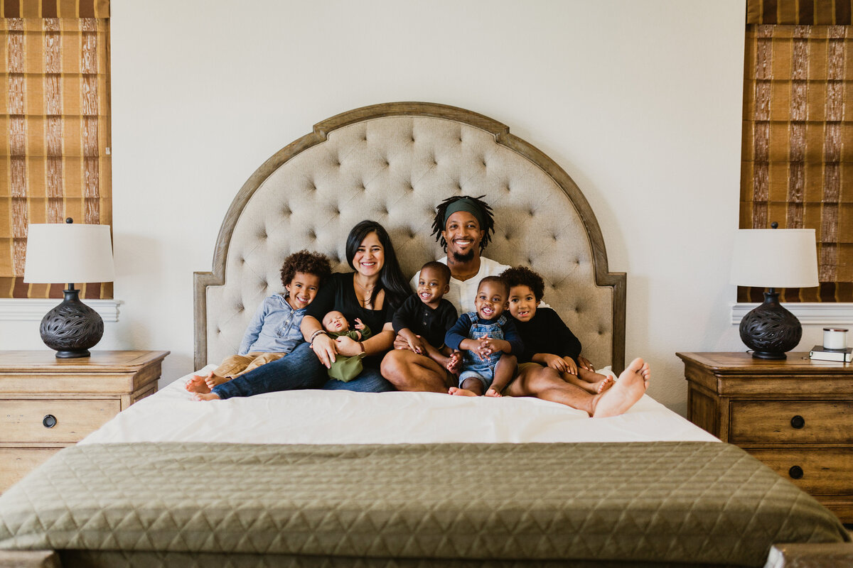 A memorable Albuquerque maternity photoshoot featuring a family of seven. The family, consisting of two adults and five children of different ages, is seated together on a white bed with green accents. All are looking at the camera, capturing a moment of unity and anticipation.