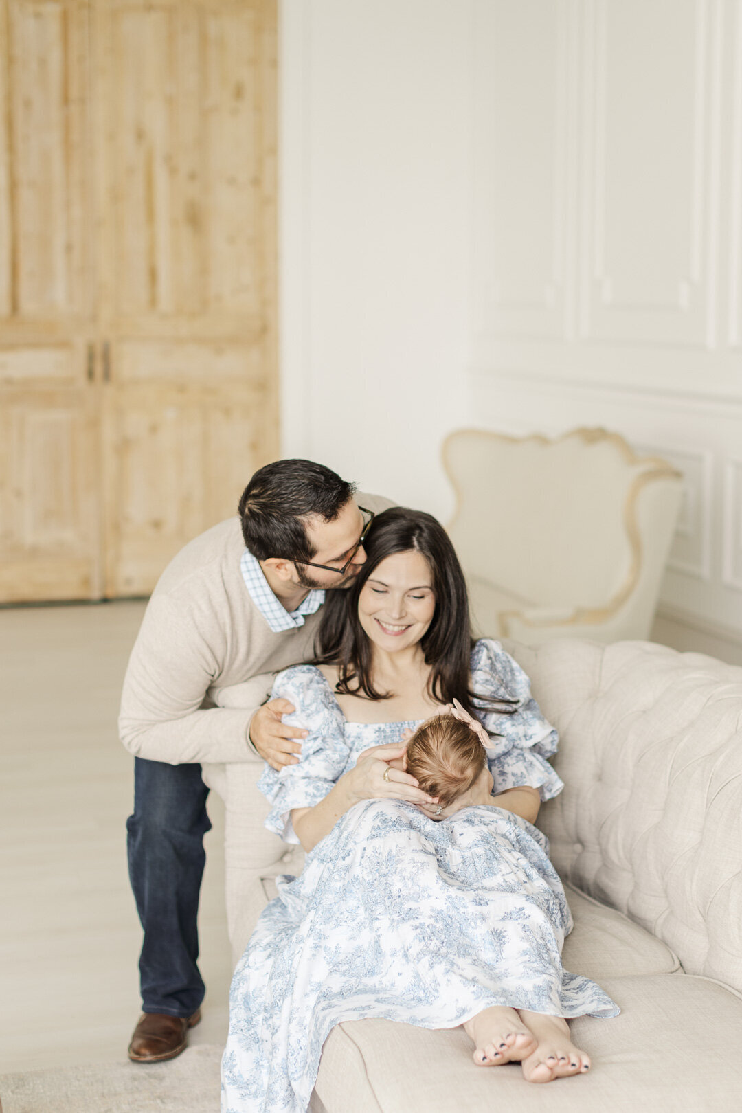 mom holds baby on sofa wearing a blue dress while dad kisses her temple for katy newborn photographer portrait session