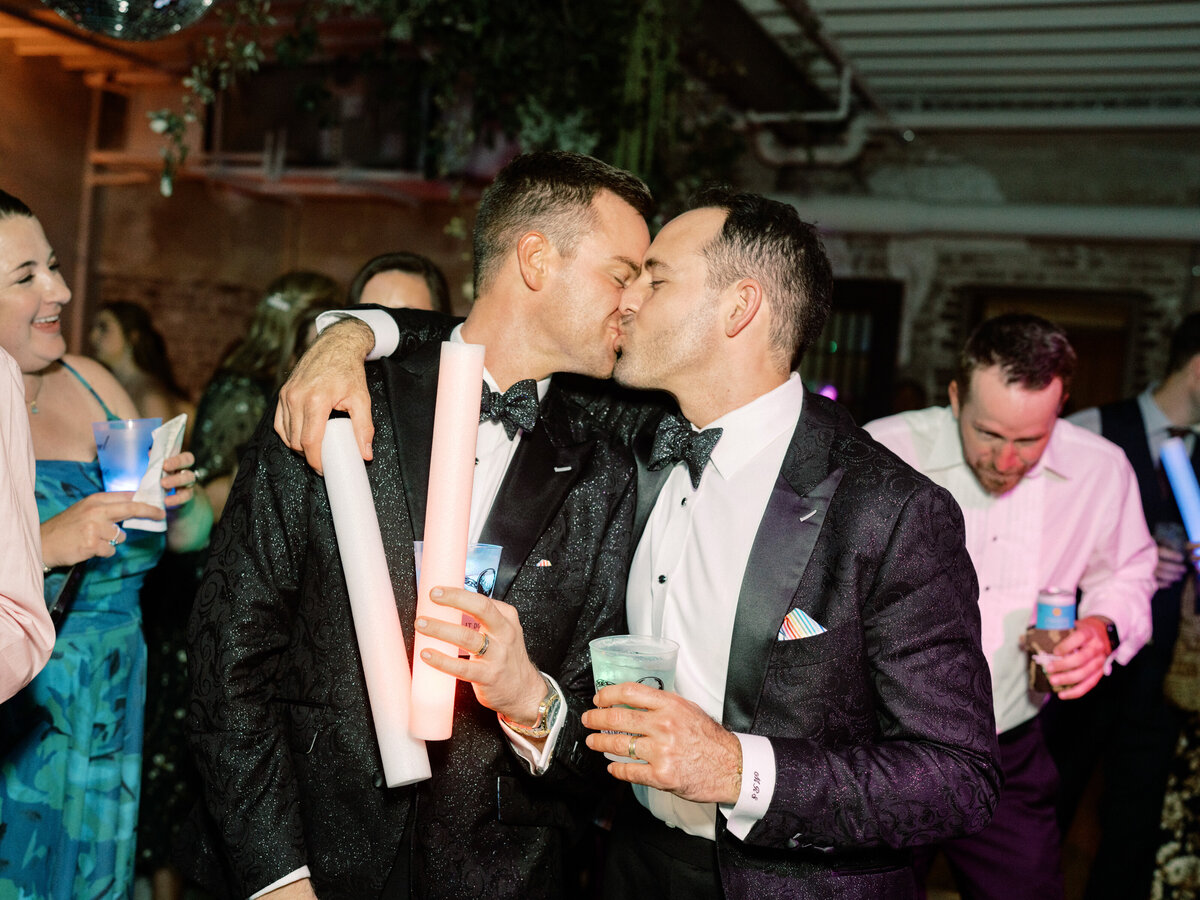 This lively image captures a heartfelt moment between two men kissing while celebrating at a festive event, highlighting love, joy, and connection in a vibrant atmosphere.