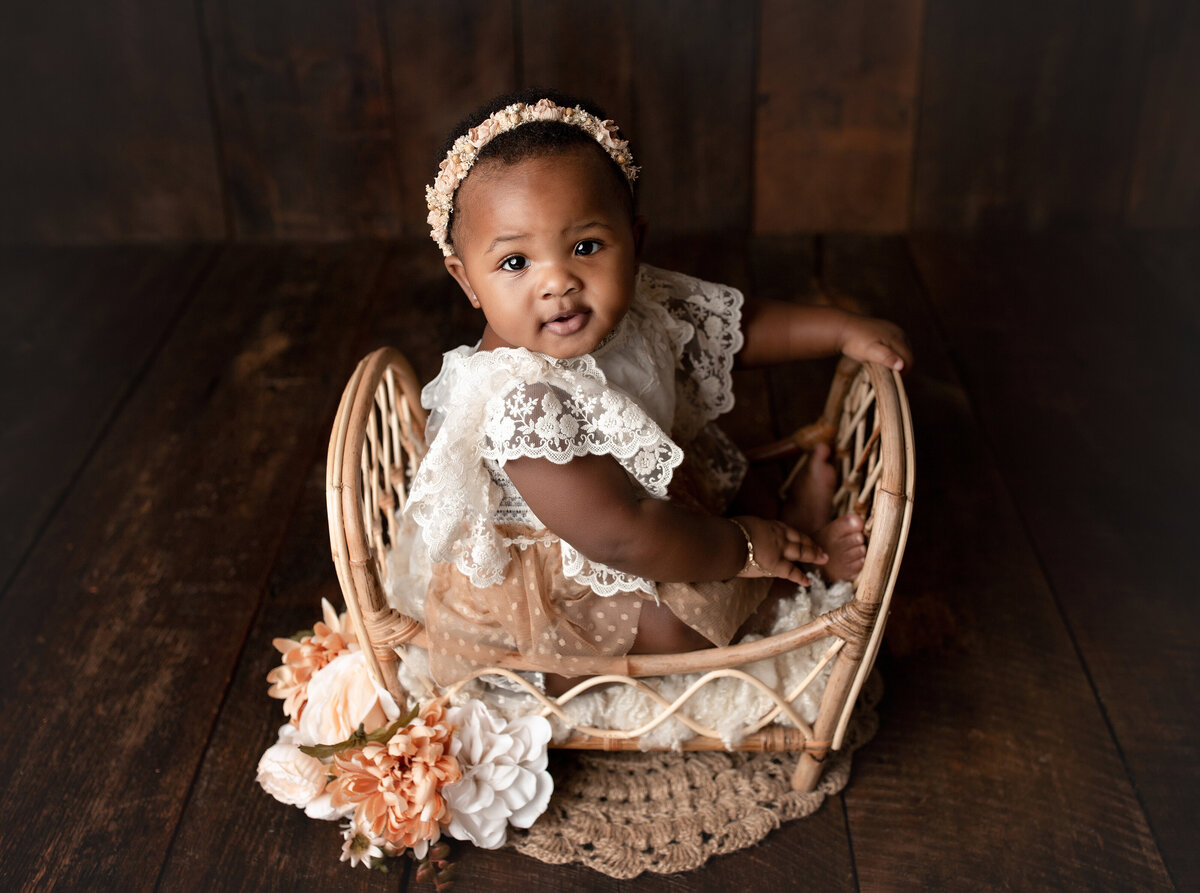 Baby girl in miniature wicker crib. Baby girl is in a lace and taupe dress and matching headband. Baby girl is looking up at the camera intently.