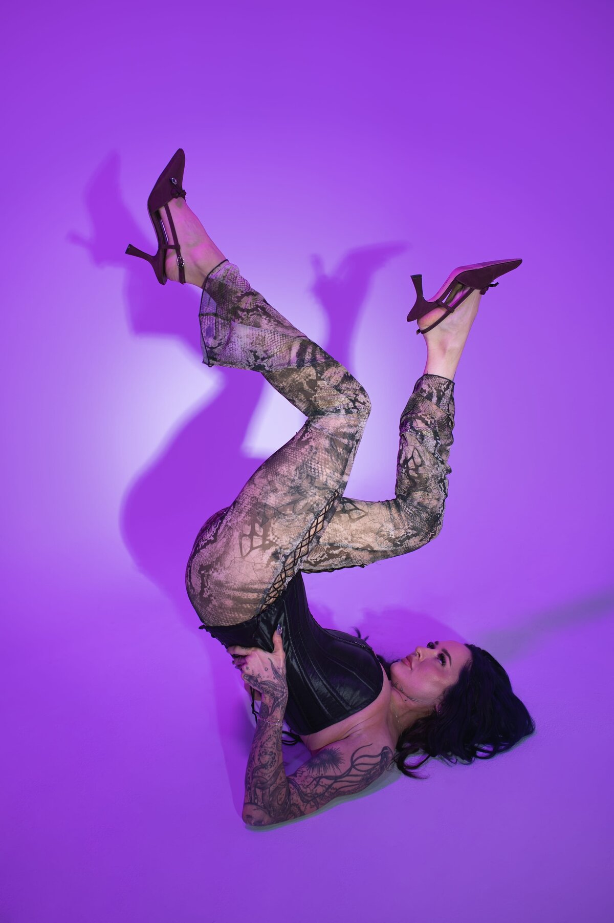 Woman with black, long hair lays in editorial pose on her back while supporting her lower back lifted in the air and her knees bent while wearing a corset, lacy pants and black kitten heels in front of a purple backdrop