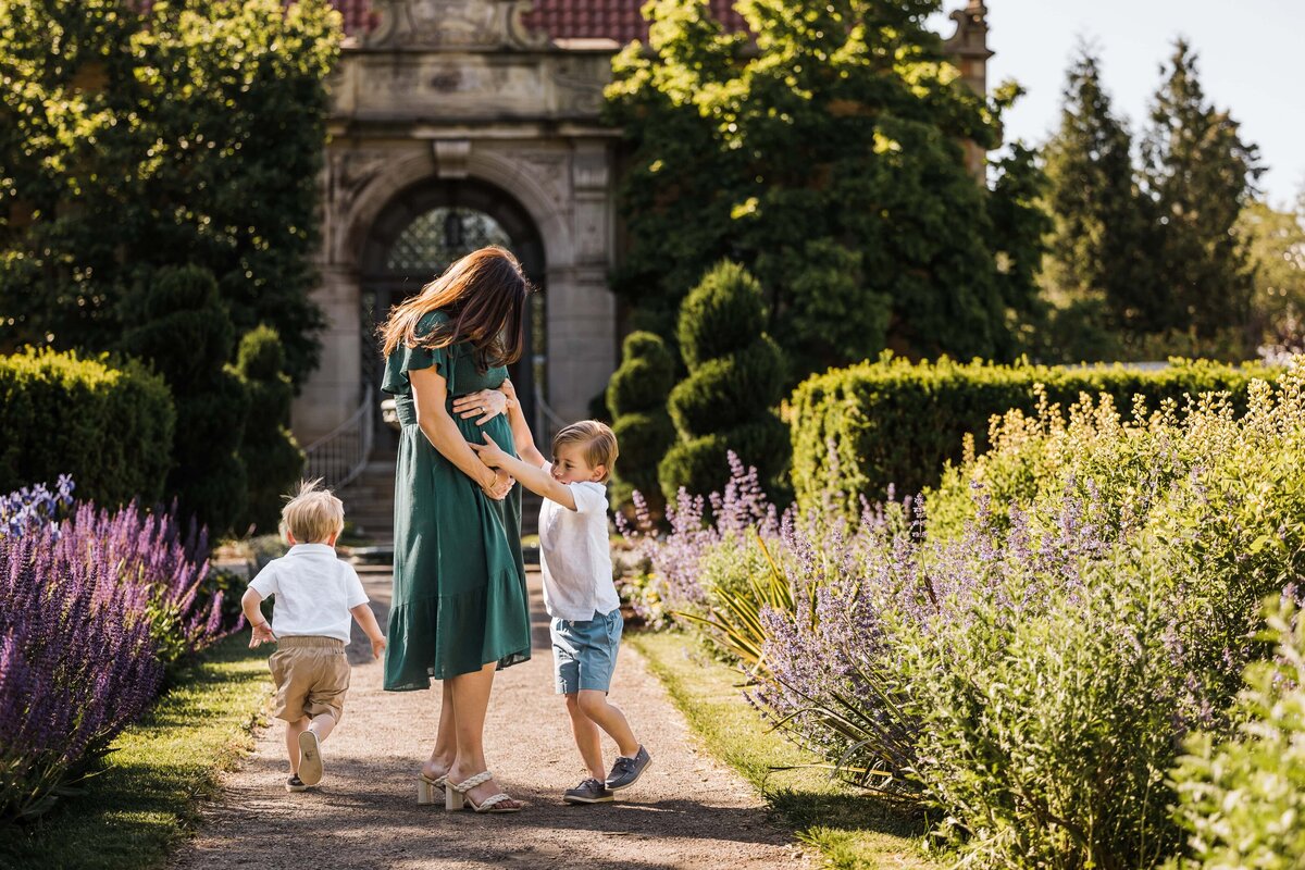 A woman holding hands with a child while walking through a garden path during a maternity photography session, with another child running ahead.