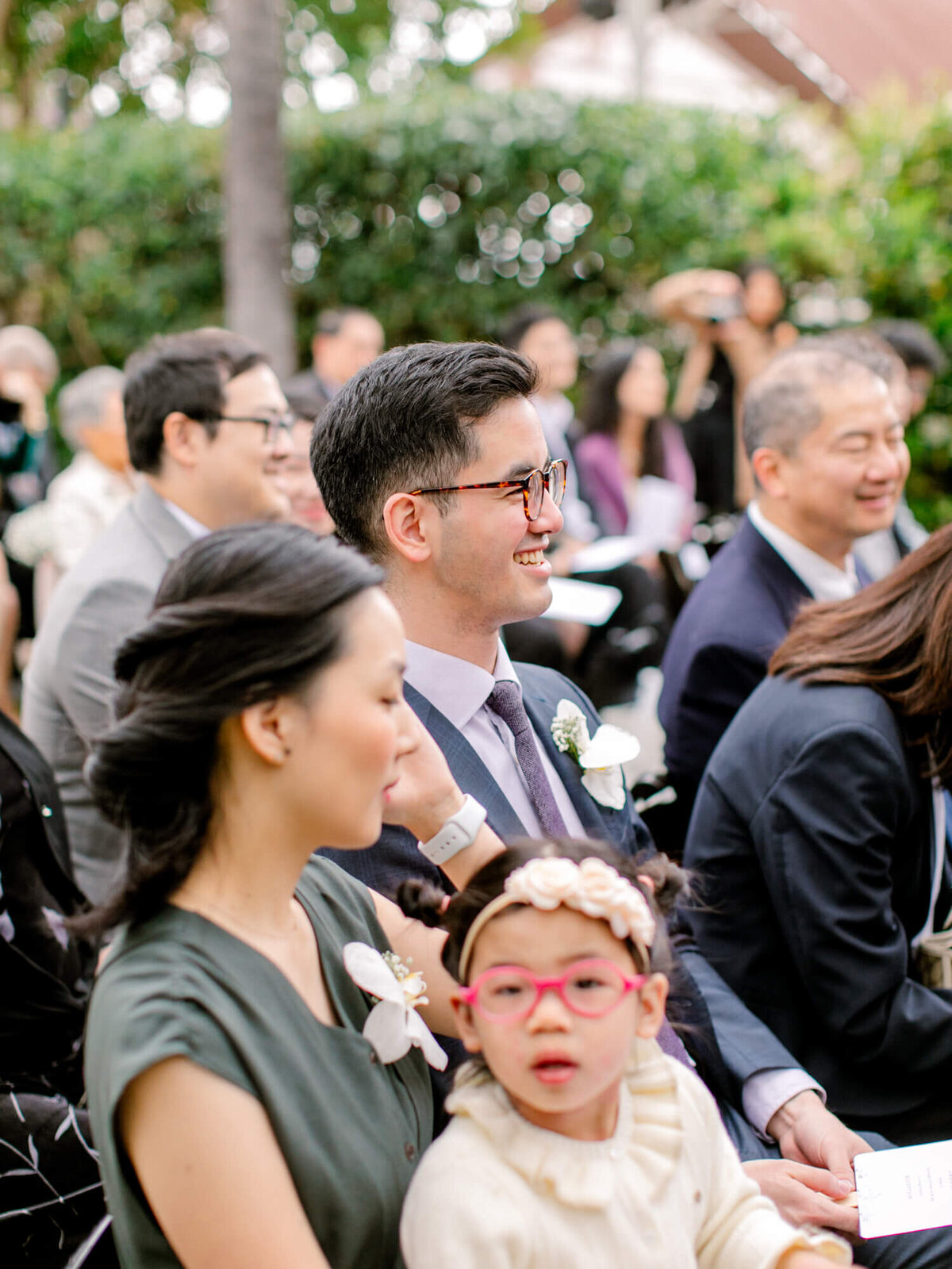 A guest in a suit and glasses smiles while seated at an outdoor event. Adults and children around him appear engaged, with greenery in the background.
