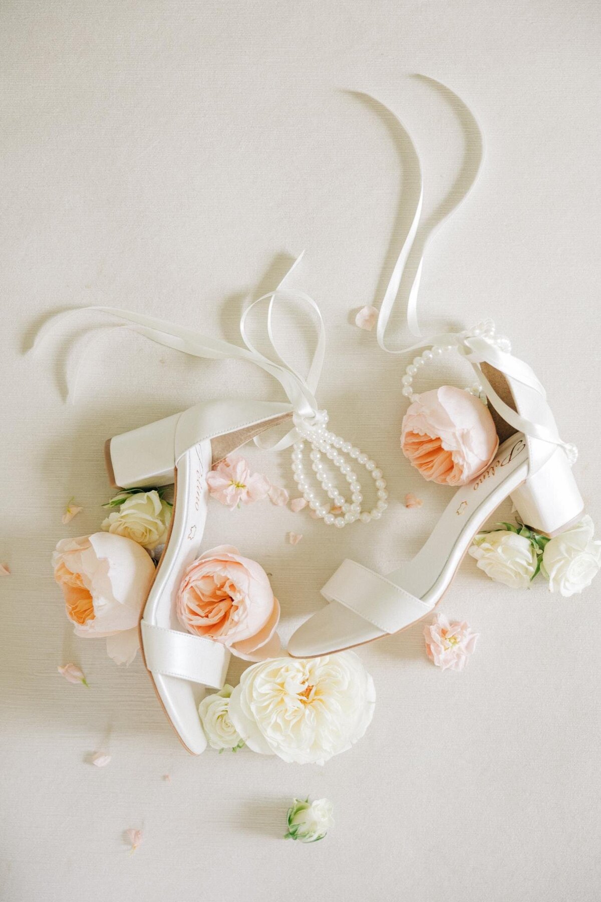 A pair of white bridal heels adorned with flowers and pearls arranged on a plain background.