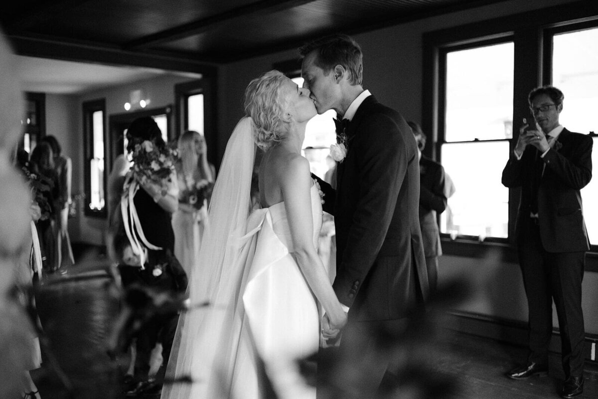 The bride and the groom kiss each other as guests take photos in Foxfire Mountain House, New York. Wedding Image by Jenny Fu Studio