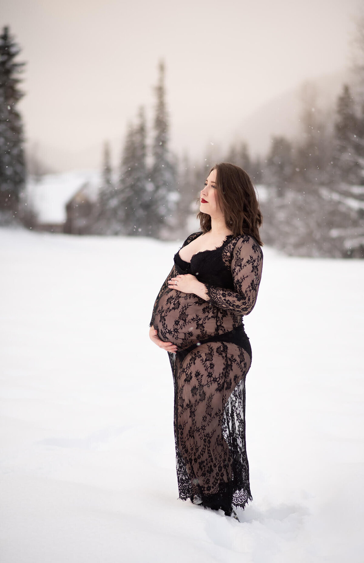 A pregnant woman wearing a black see through lace dress caresses her belly while standing in the snow in this beautiful winter backdrop at Sunpeaks, BC. Taken by Brittany Danielle Photography