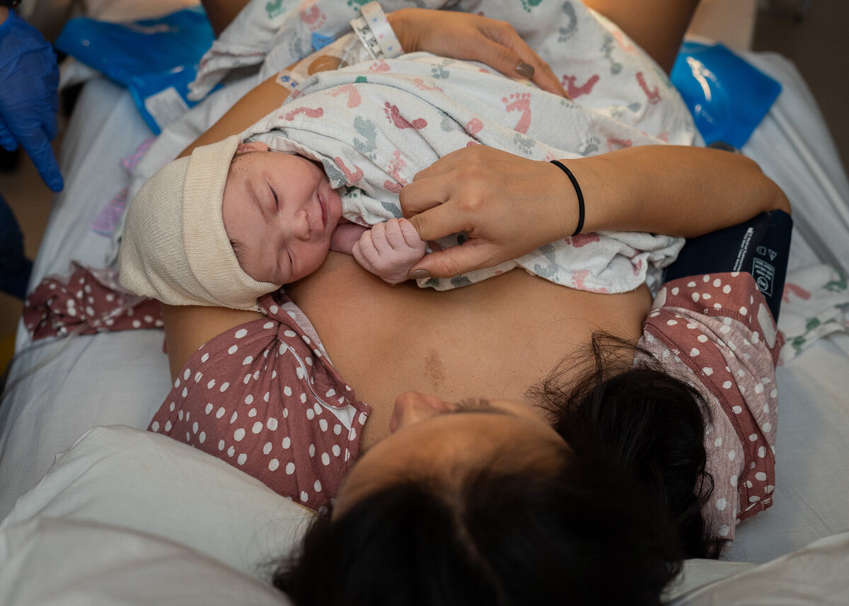 A newborn baby is laying on it's mother's chest at a hospital in Seattle, WA. The mother is holding the tiny baby's hand.