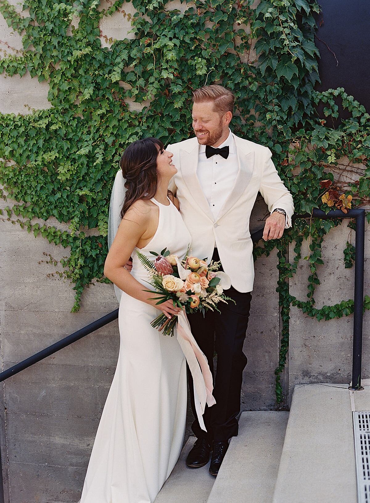 The bride, wearing a silk sheath halter top wedding dress and veil, holding a large bouquet of pink, peach, blush, ivory and white flowers with ferns and palm fronts as she smiles up at the groom who is wearing a tuxedo with a white jacket . The bride and groom are lounging in the ivy covered courtyard of Clementine Hall against a soft gray wall.