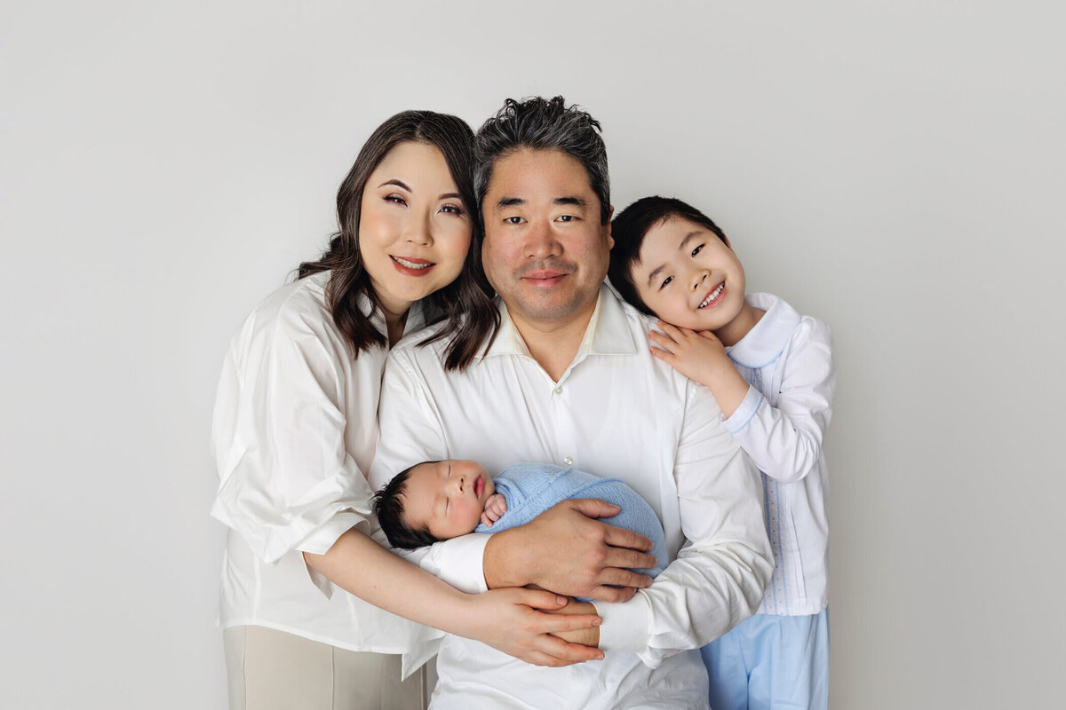 A family portrait featuring a mother, father, and two children against a white background. The mother and father, both wearing white tops, hold a sleeping newborn swaddled in a blue blanket. The older child stands next to the father, smiling and leaning affectionately on his shoulder. The family exudes warmth and happiness.