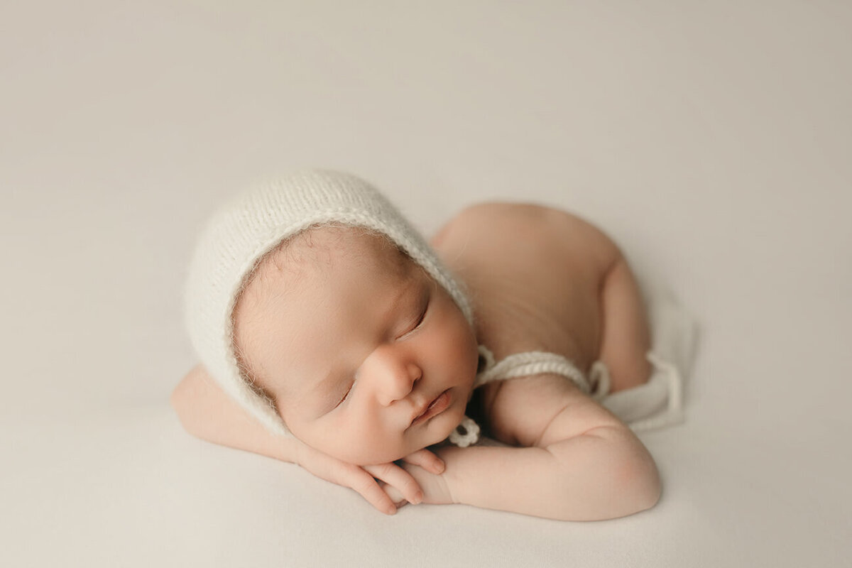boy sleeping on white drop with chin on hands with a white bonnet