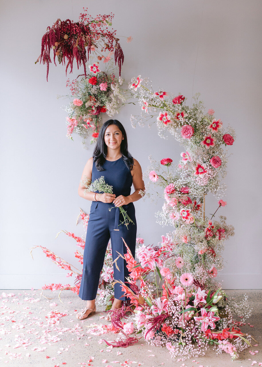 Kathiya standing in front of a dramatic pink and red floral installation, holding a bouquet and smiling