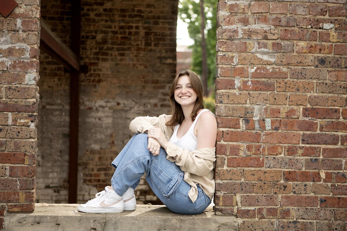 A high school senior smiles brightly in front of a classic brick wall for a memorable photo session