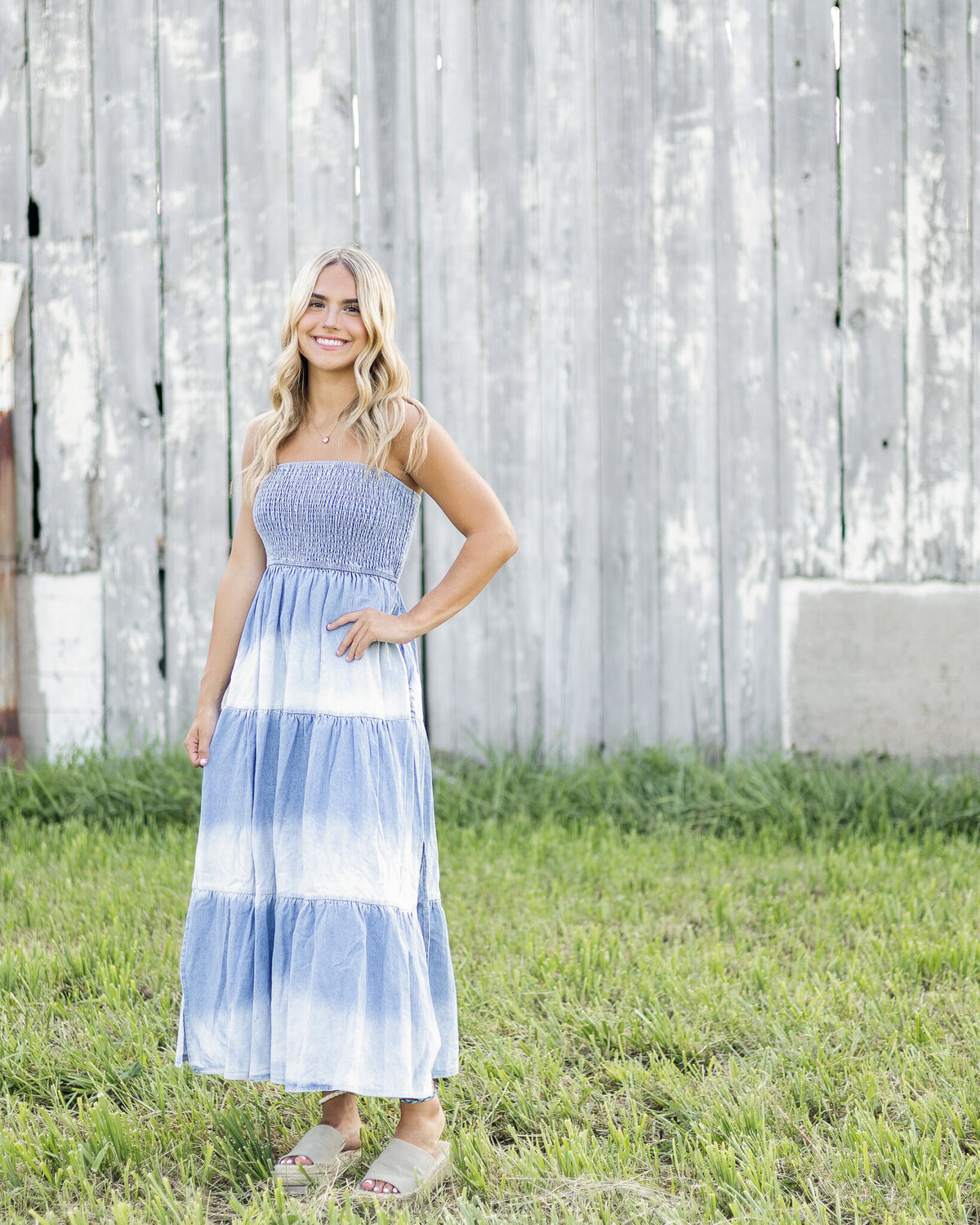high school senior female, standing standing in grass in front of a gray and white barn, with long blonde hair, wearing a white and blue sleeveless dress, smiling at the camera with one hand on her hip.