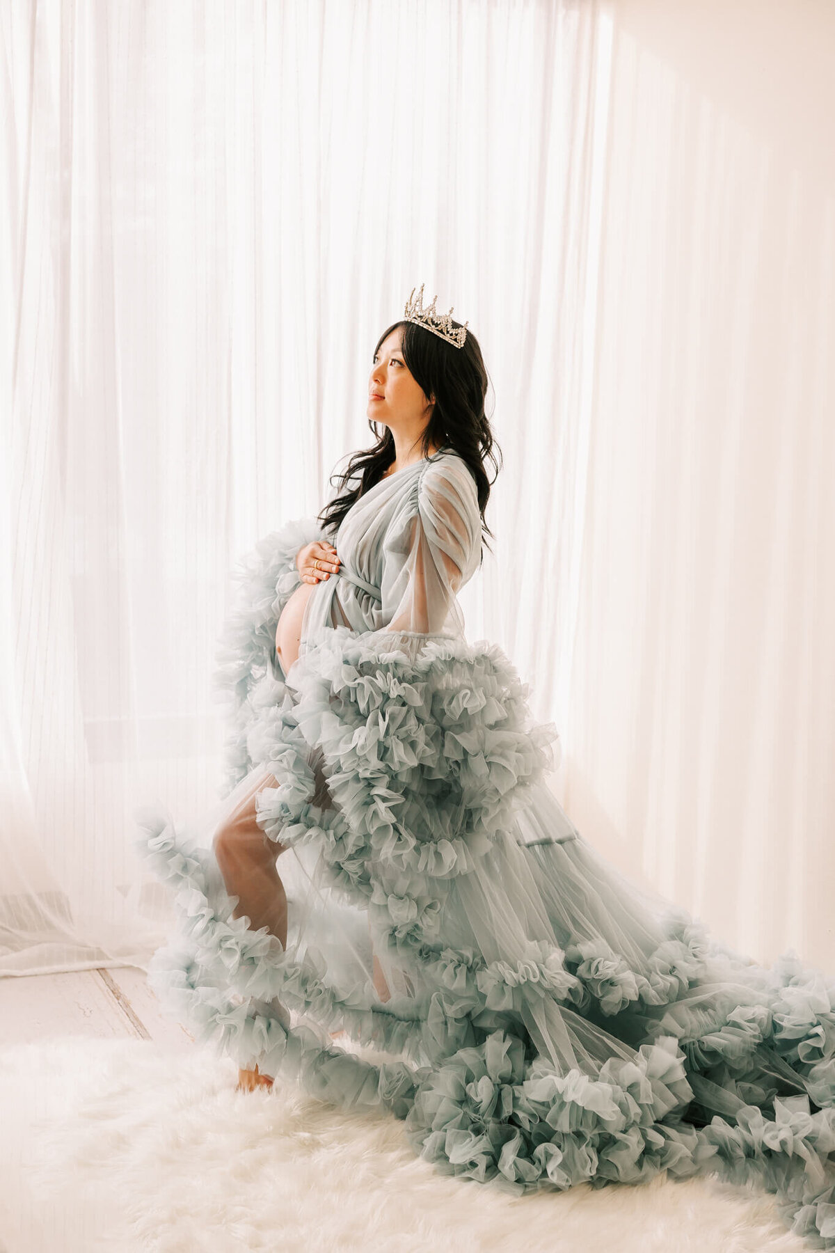 pregnant woman standing in front of window with her belly showing and crown on her head. She is looking off to the left with the couture gown flowing behind her.
