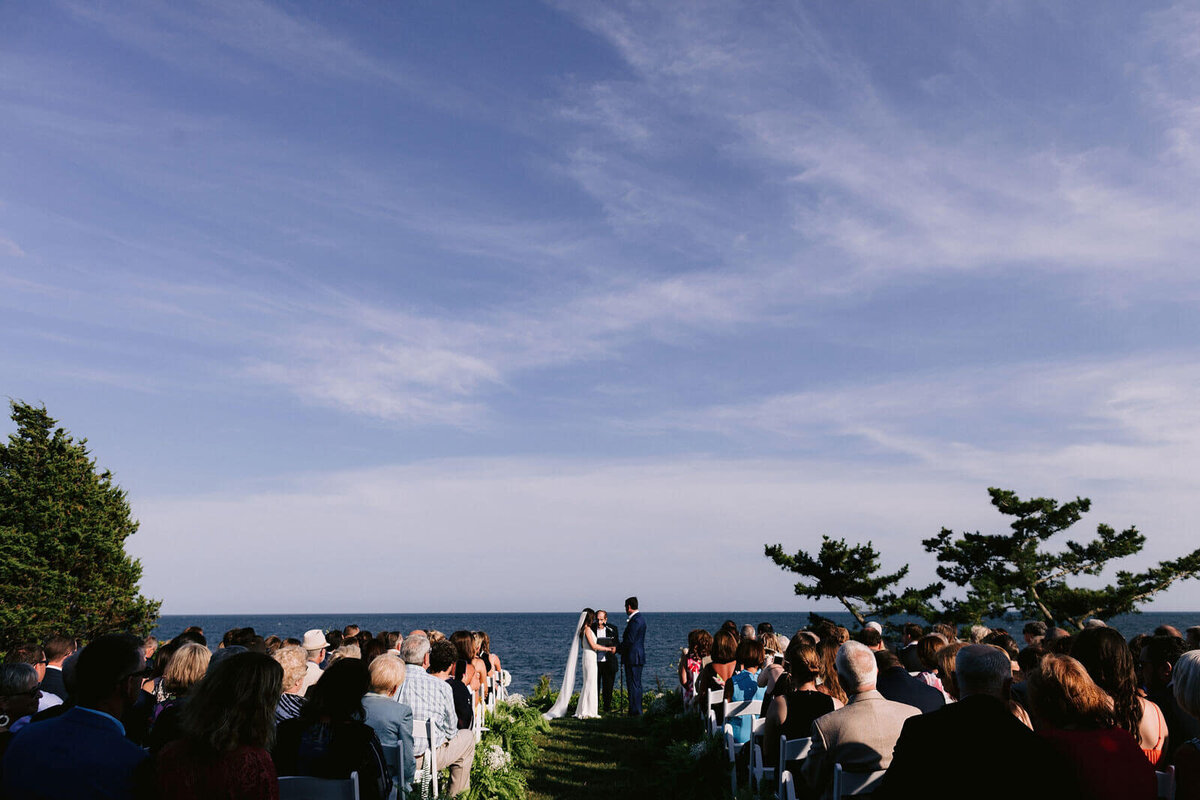 A wedding ceremony is being held in Wianno, Cape Cod, MA, with the sea and vast blue sky in the background.