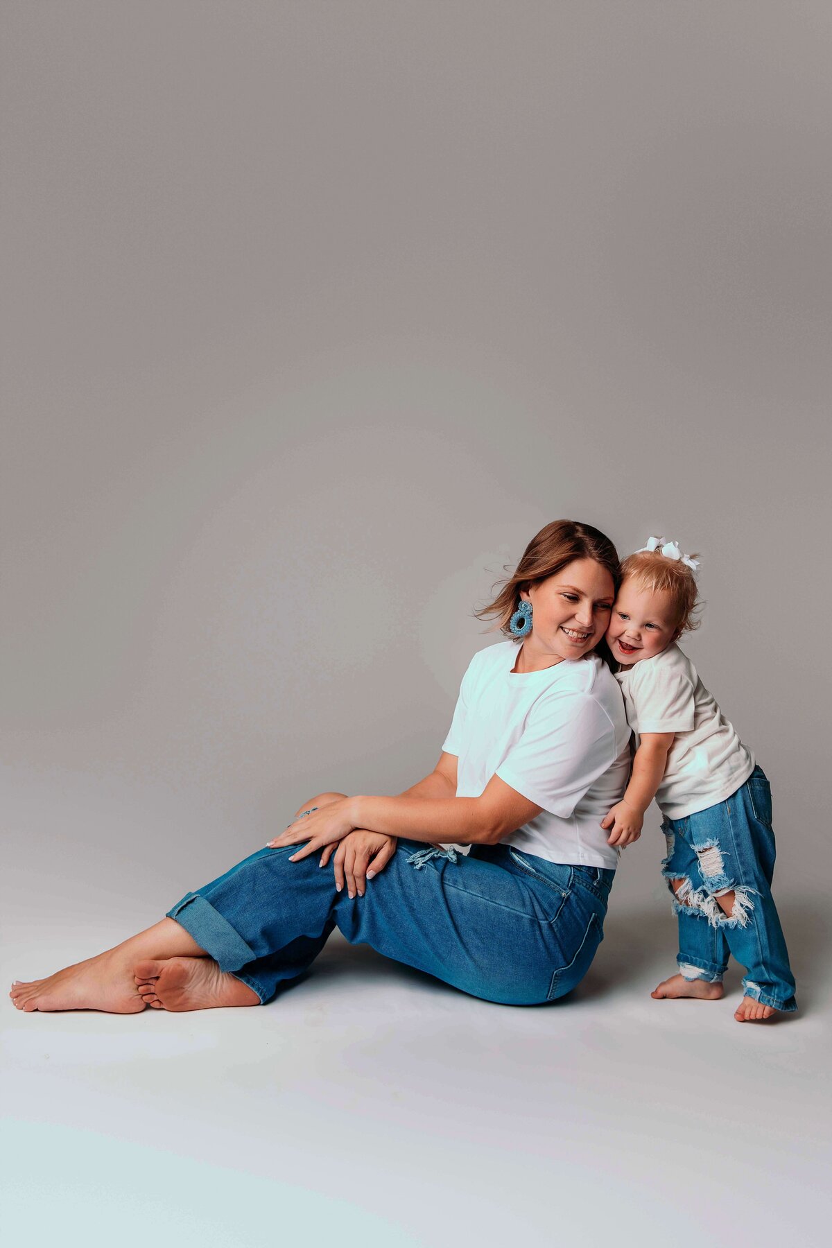 A young mom and her toddler daughter pose for a playful portrait by Mobile, AL Motherhood Photographer. They are wearing matching jeans and shirts, and are barefoot. The photo shows a happy duo, as the child stands behind her sitting mom. The child is leaning against her mother's back for a quick embrace, as her mother looks lovingly at her.