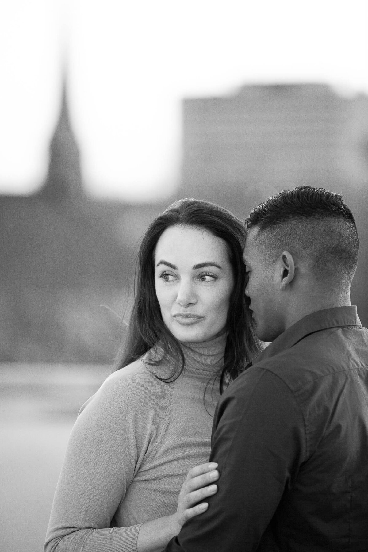 Woman looking to the side. Man is holding her close with head at her temple. Blurry Charleston city in background. Charleston engagament photographer