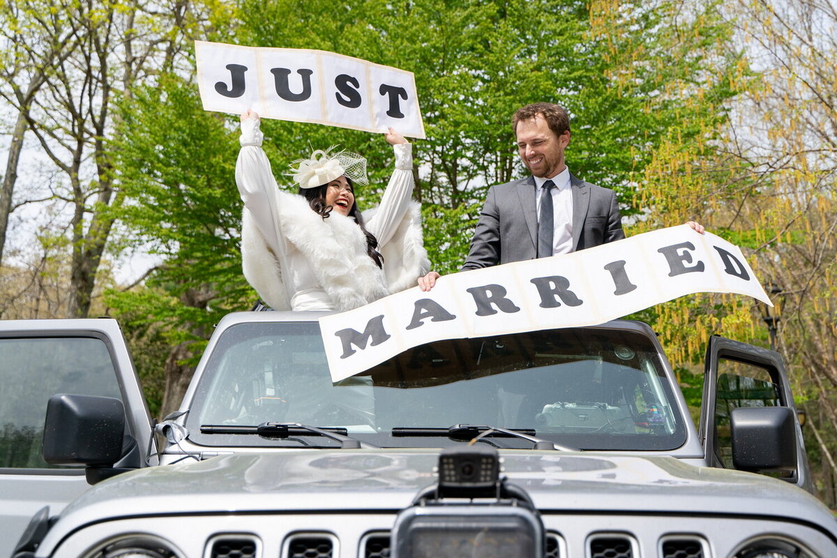 Bride and groom pop out of the sun roof of a silver SUV, proudly showing DIY Just Married signs
