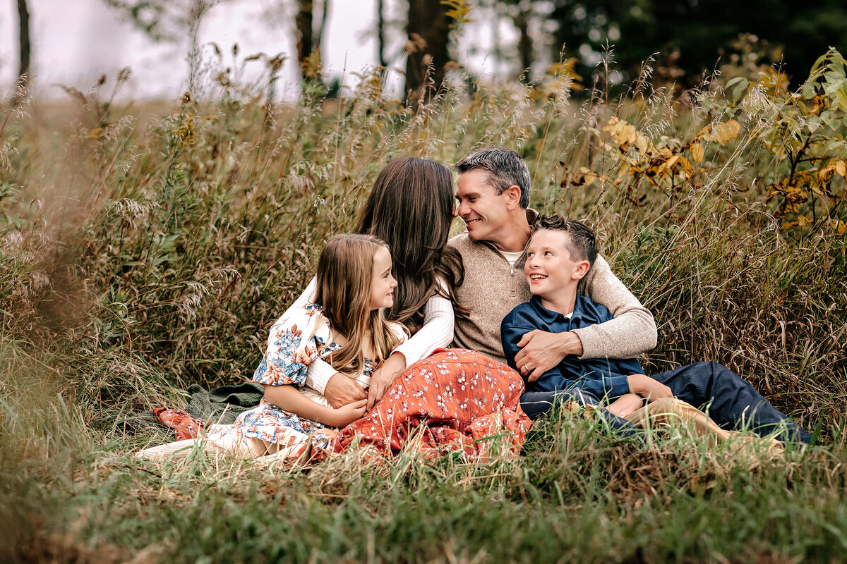 A family of four sits together in a field with tall grass and trees. The parents and two children, a girl and a boy, are embraced in a warm hug, smiling at each other. They're casually dressed, surrounded by autumn foliage.
