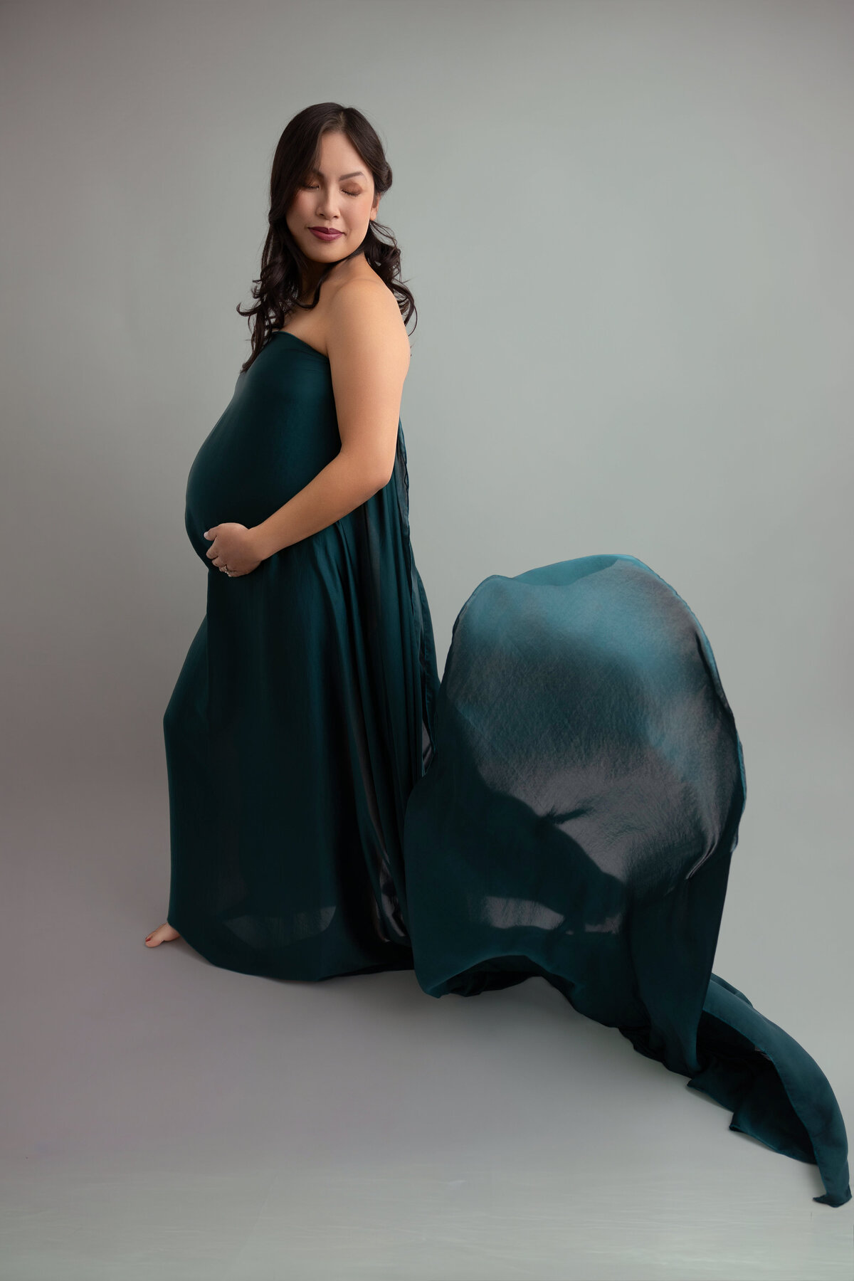 A pregnant mother wearing a silk forest green fabric posed against a light green backdrop