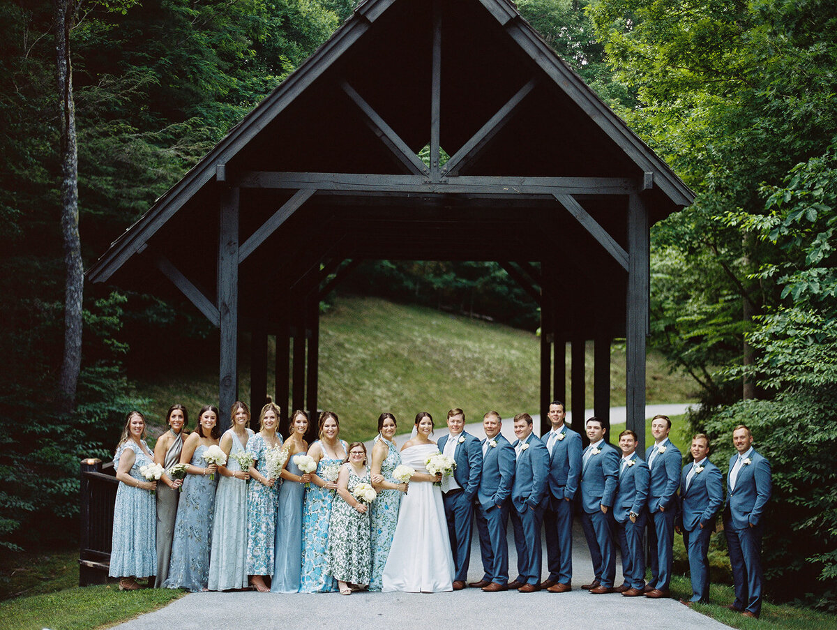 Full bridal party portrait on a rustic covered bridge surrounded by lush greenery at a Cashiers, NC mountain wedding.
