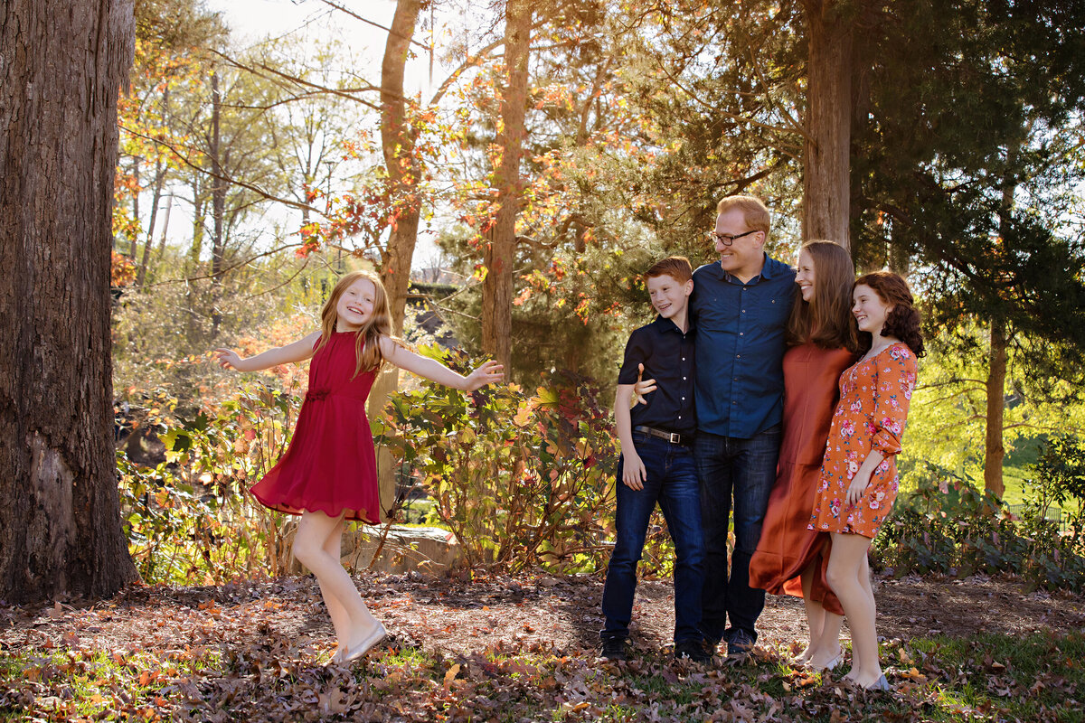 Endearing scene of red‑haired girl twirling in golden leaves while her family embraces and smiles in a wooded park at Ballantyne Corporate Park Charlotte NC