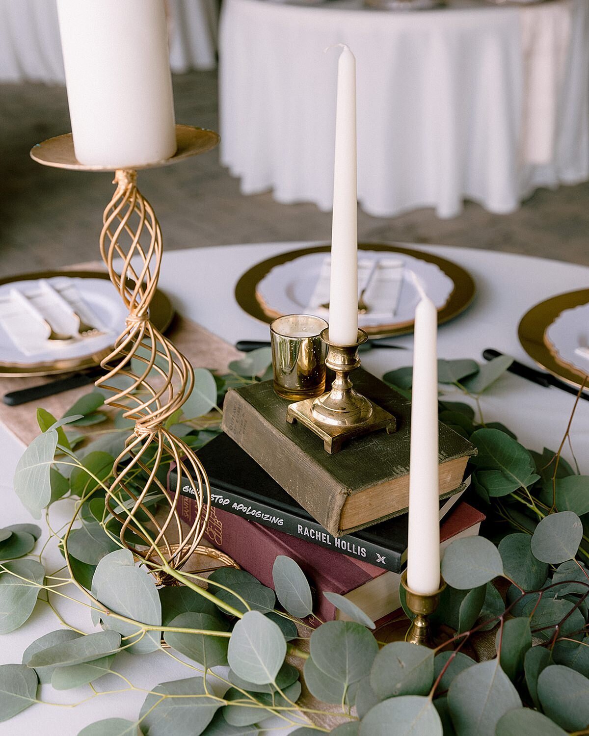 Stacks of vintage books in the center of each table topped with gold candle sticks and white table candles surrounded by silver dollar eucalyptus at a wedding reception at Ravenswood Mansion. The tables are set with gold chargers, white plates and white flatware is set at each place setting.