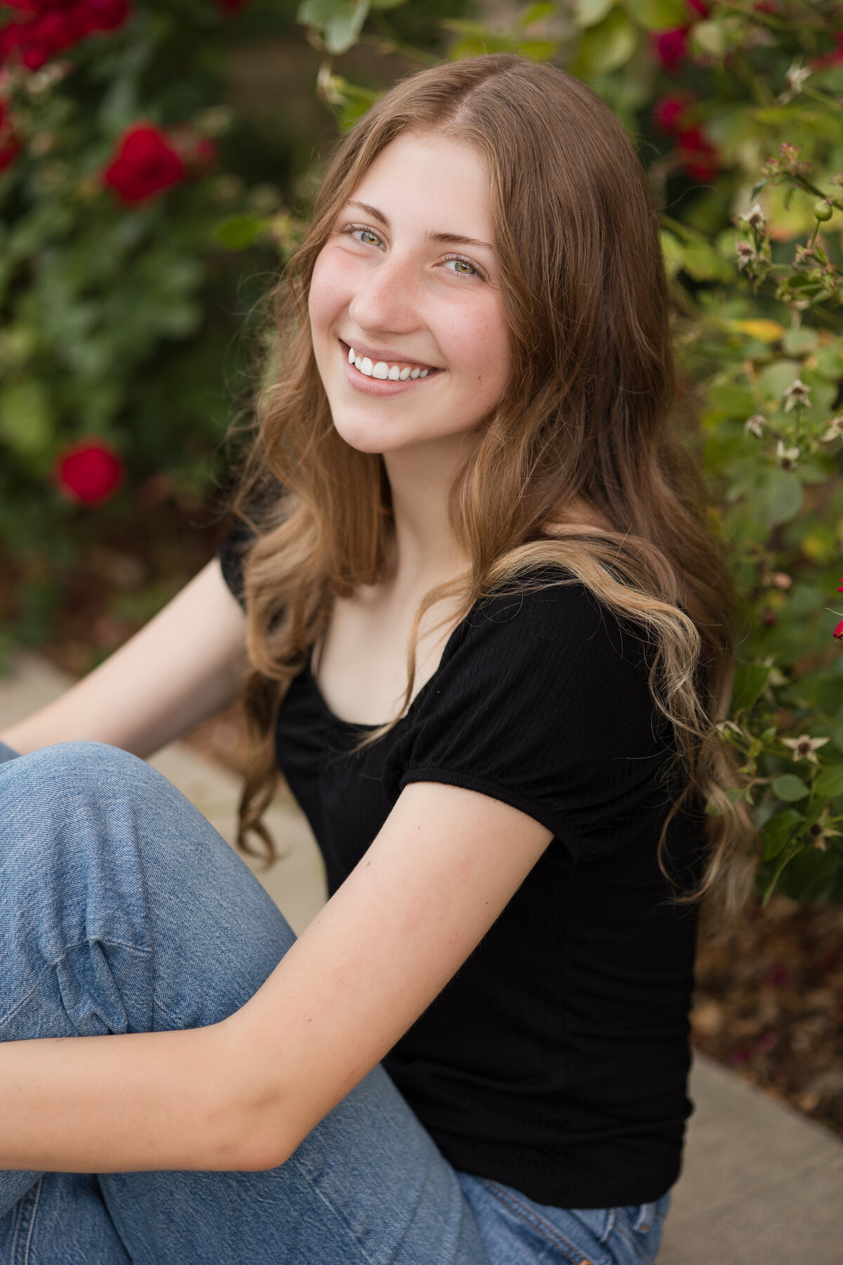 Senior girl seated in front of a rose bush smiling at the camera