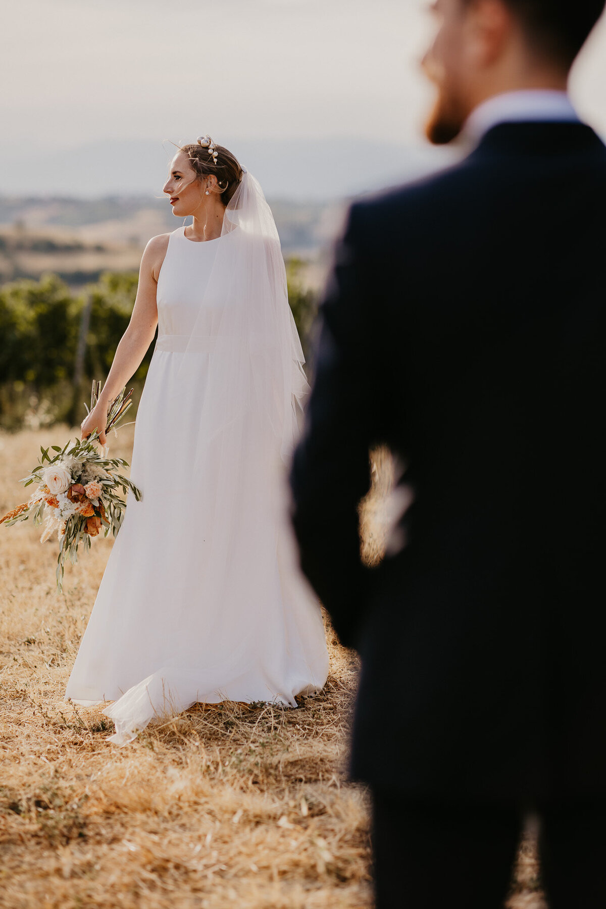 Bride smiling at sunset with groom in background, vineyards near Villa Dianella, Tuscany wedding photographer.