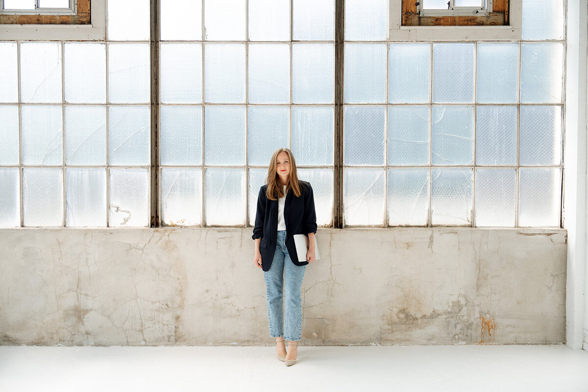 Modern Orange County branding portrait of a woman holding a laptop, standing in front of textured industrial windows.