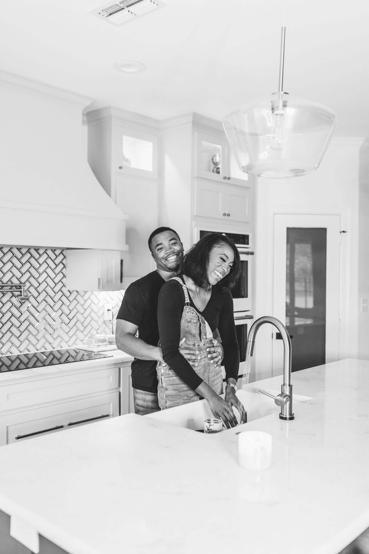 black and white photo of husband and wife laughing in kitchen while washing dishes during in home family lifestyle photography seeesion