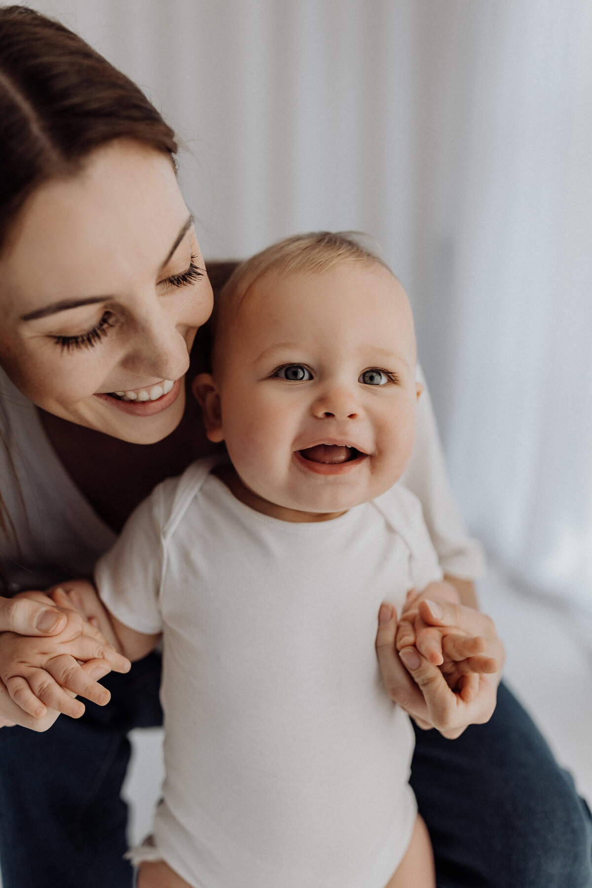 A mother holding her little baby who is wearing a white top to match hers and laughing towards the camera during a baby milestone photoshoot