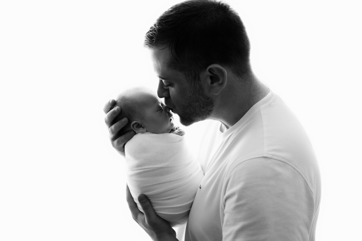 Dad wearing white tenderly kissing his newborn on the nose on a bright white background.