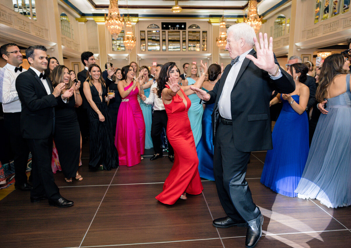 A lively ballroom scene with a group of elegantly dressed people. A woman in a red dress dances energetically with an older man in a suit. The crowd, wearing colorful evening gowns and suits, claps and smiles under a chandelier-lit ceiling.