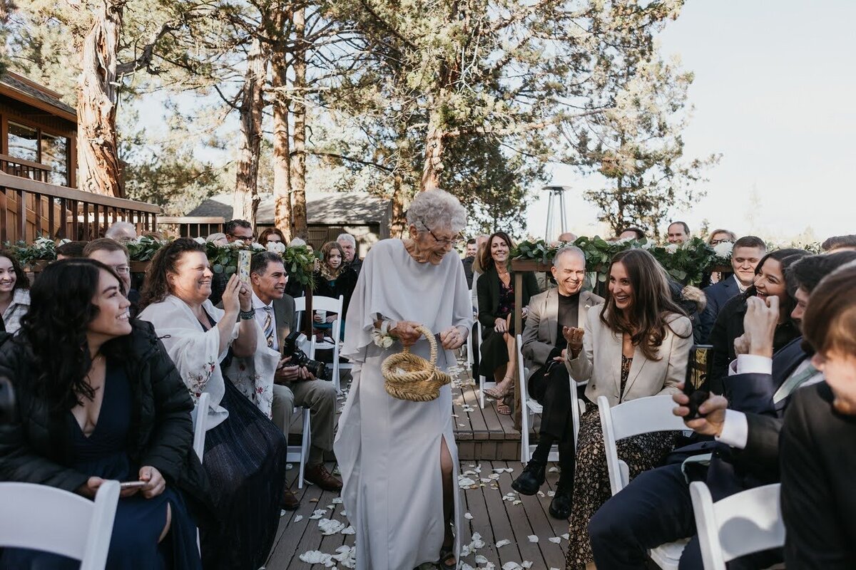 Flower grandma carries a basket of flower petals of the wedding aisle as guests look on and smile
