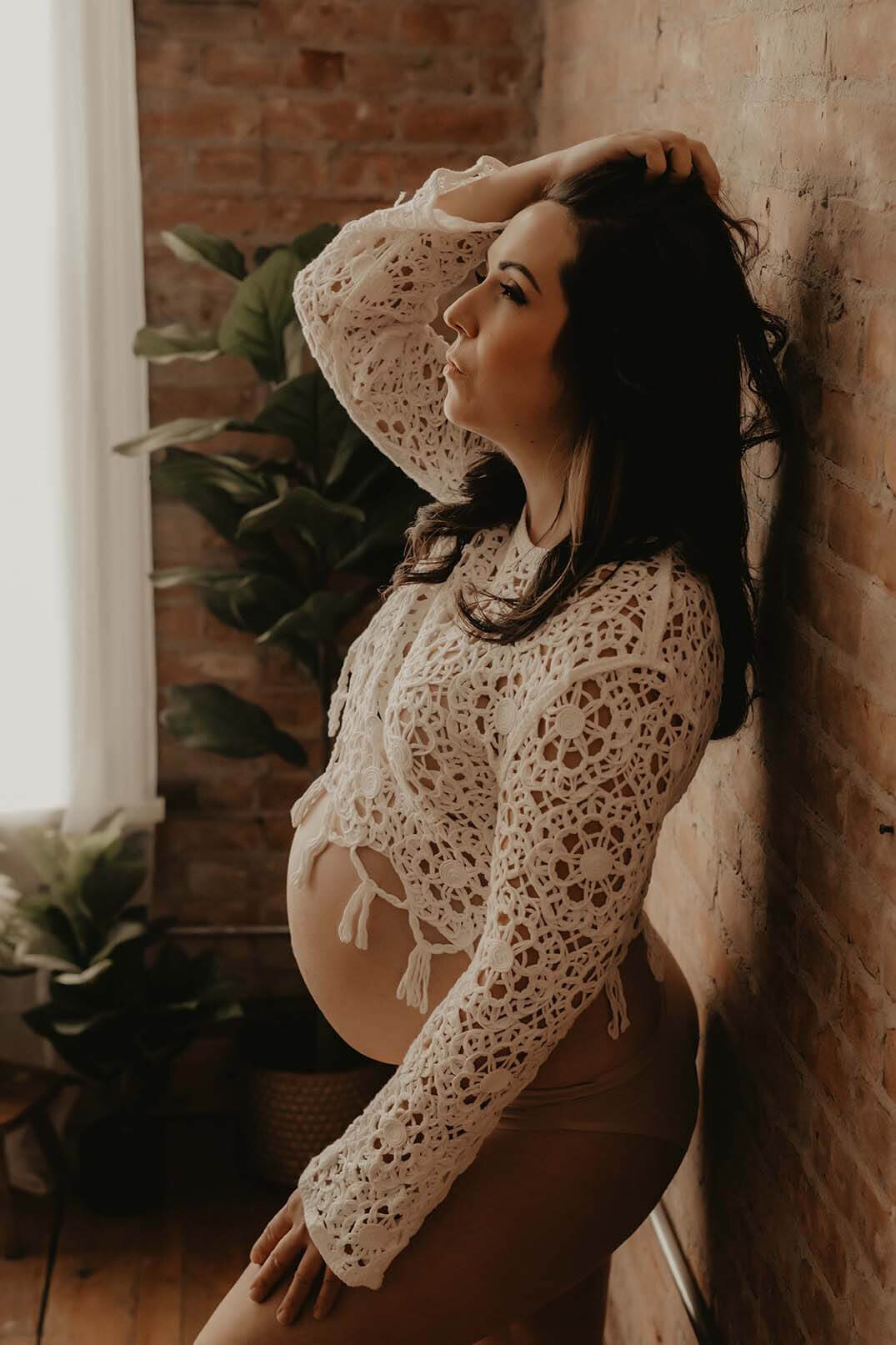 A beautidul pregnant woman holds her hair while resting her other hand on her thigh against a brick wall in a beautifully lit studio in Canandaigua NY