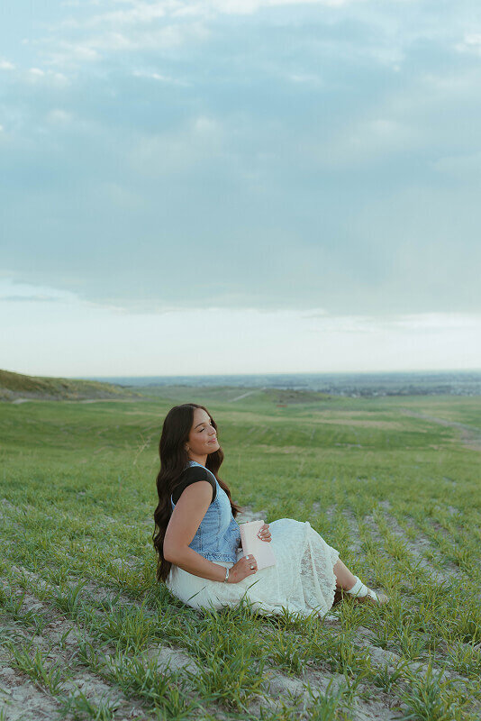 A woman sits in a grass field as she holds her Book of Mormon. She's looking to the side.