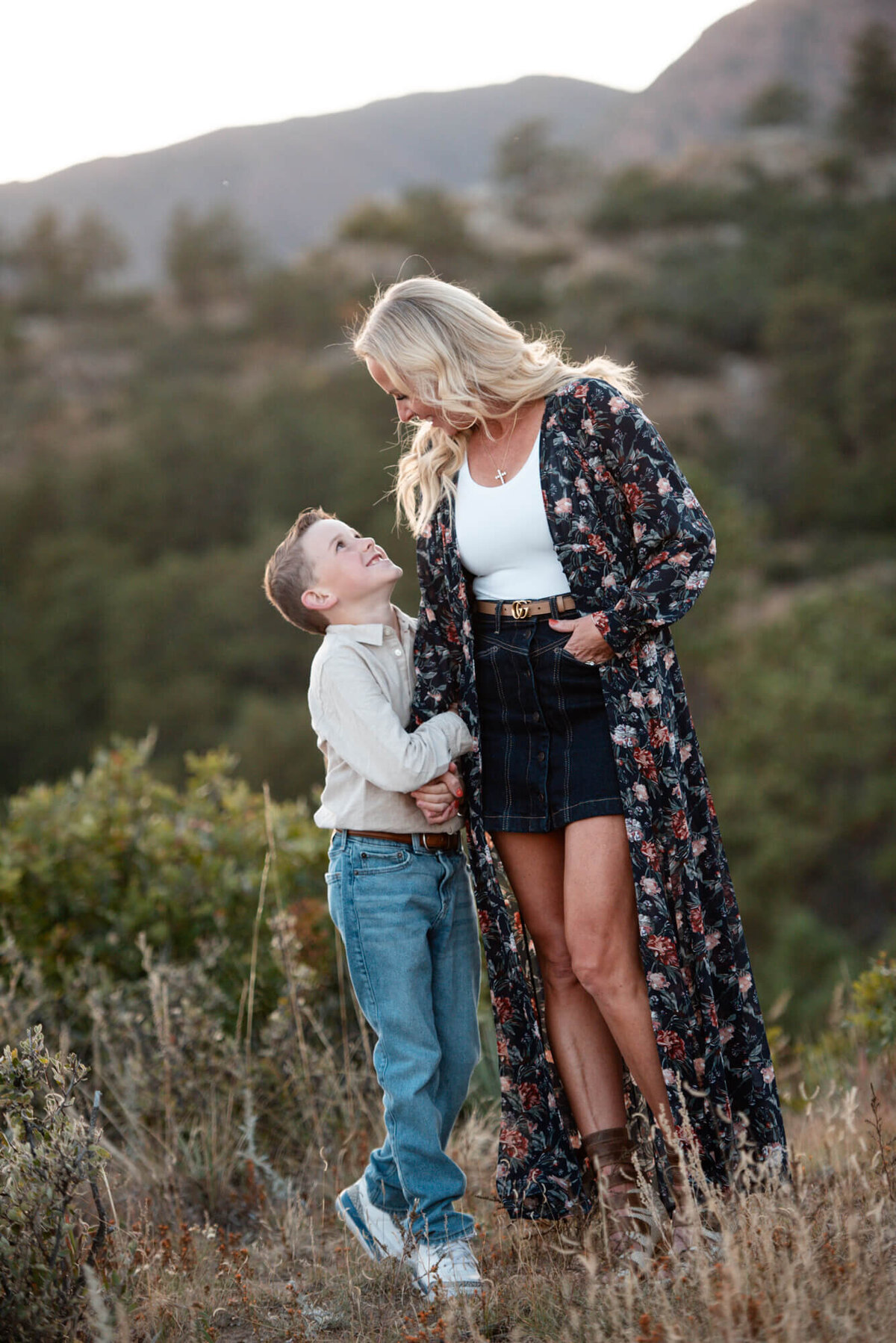 A young boy holds his mom's hand while smiling up at her while standing in tall grass