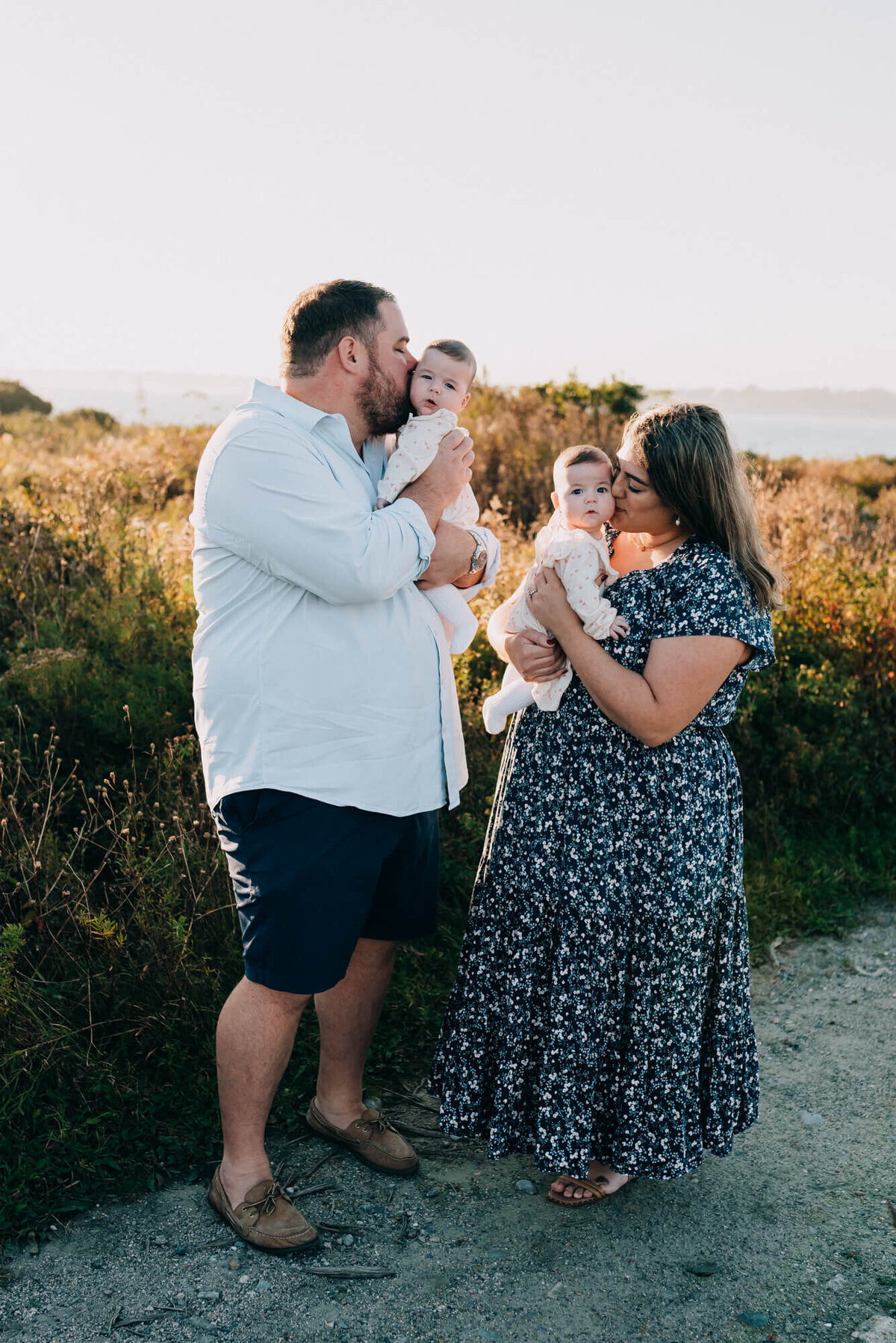 A mother and father stand next to the ocean at sunset holding and kissing their young twin daughters