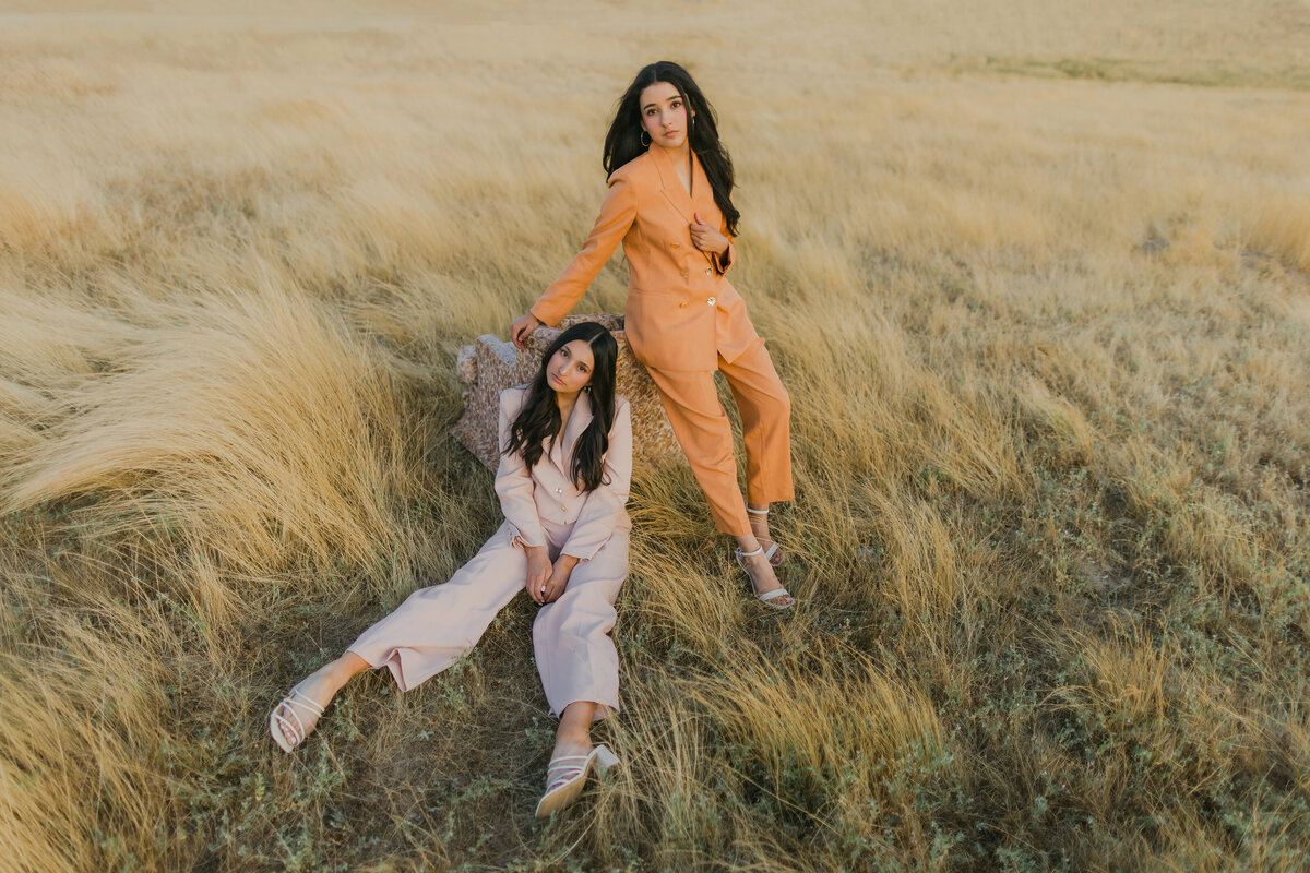 Two women pose in pastel and orange suits in a golden field—one standing and one seated on vintage upholstery—evoking sisterhood and softness. Portrait photography in Greenville South Carolina by Dani Lacey Photographs.