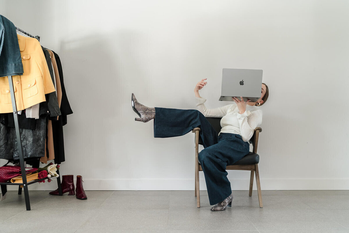 woman stylis sitting in a chair with clothing rack and holding a laptop in front of her face 