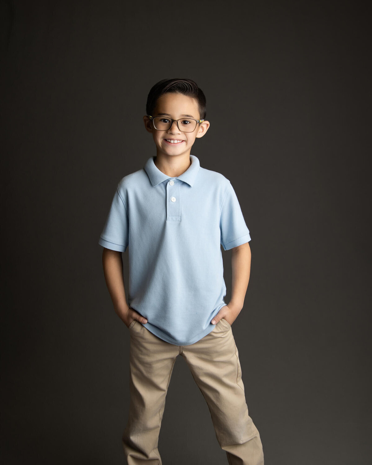 Boy with glasses posing for a school portrait in a light blue uniform shirt and khaki pants