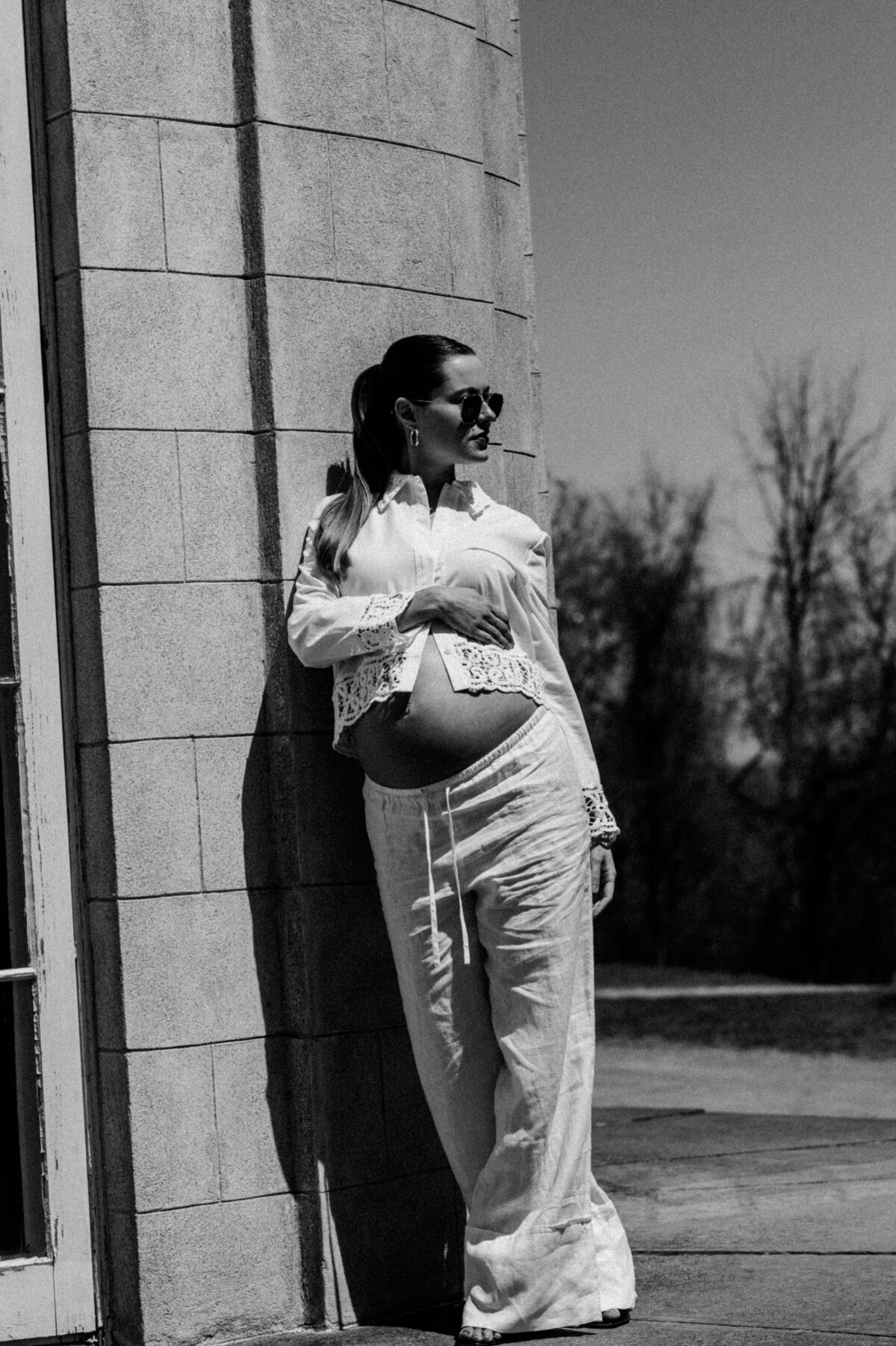 Candid black-and-white fashion-inspired maternity shot with the subject leaning against a sunlit exterior wall

