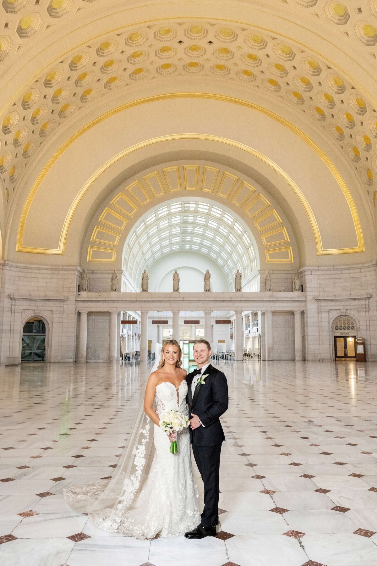 A bride in a white gown and a groom in a black tuxedo stand together in a grand hall with a high-arched ceiling and ornate architectural details. The bride holds a bouquet of white flowers.