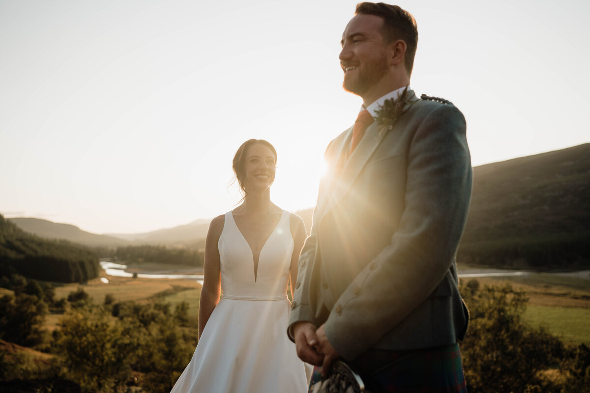 A wide photograph of a couple standing beneath the golden hour sun. The photo is warm and moody and the Mar Lodge valley can be seen behind them.