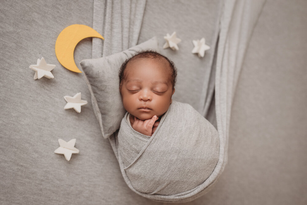 Newborn photos with Atlanta, Georgia newborn photographer of sleeping baby boy with hands under chin wrapped in gray swaddle in gray fabric swing and puff ball gray hat with stars, clouds and moon felt pieces laying beside him