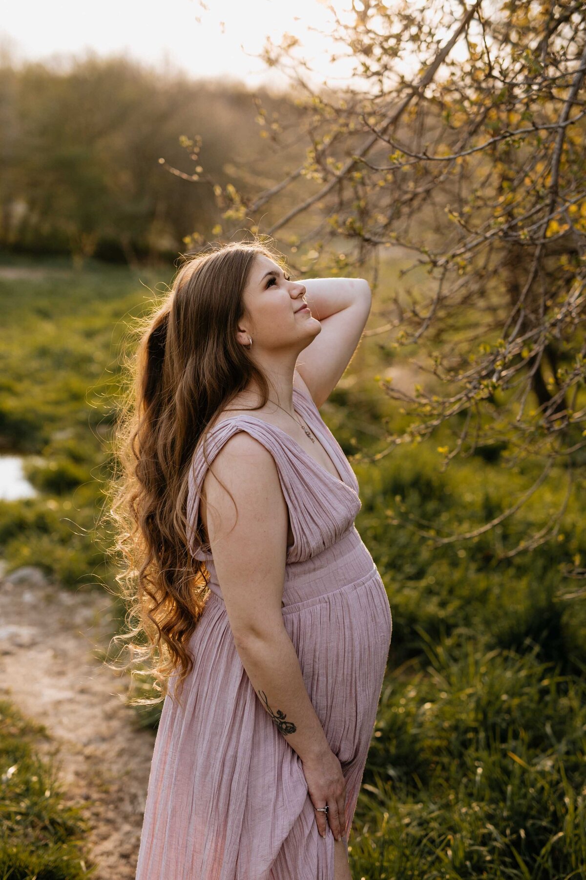 Maternity photo session. Mom in London, Ontario field wearing a pink gown. Mom is looking up at the sky and her forehand under her bump. Her back hand is brushing her hair out of her face.