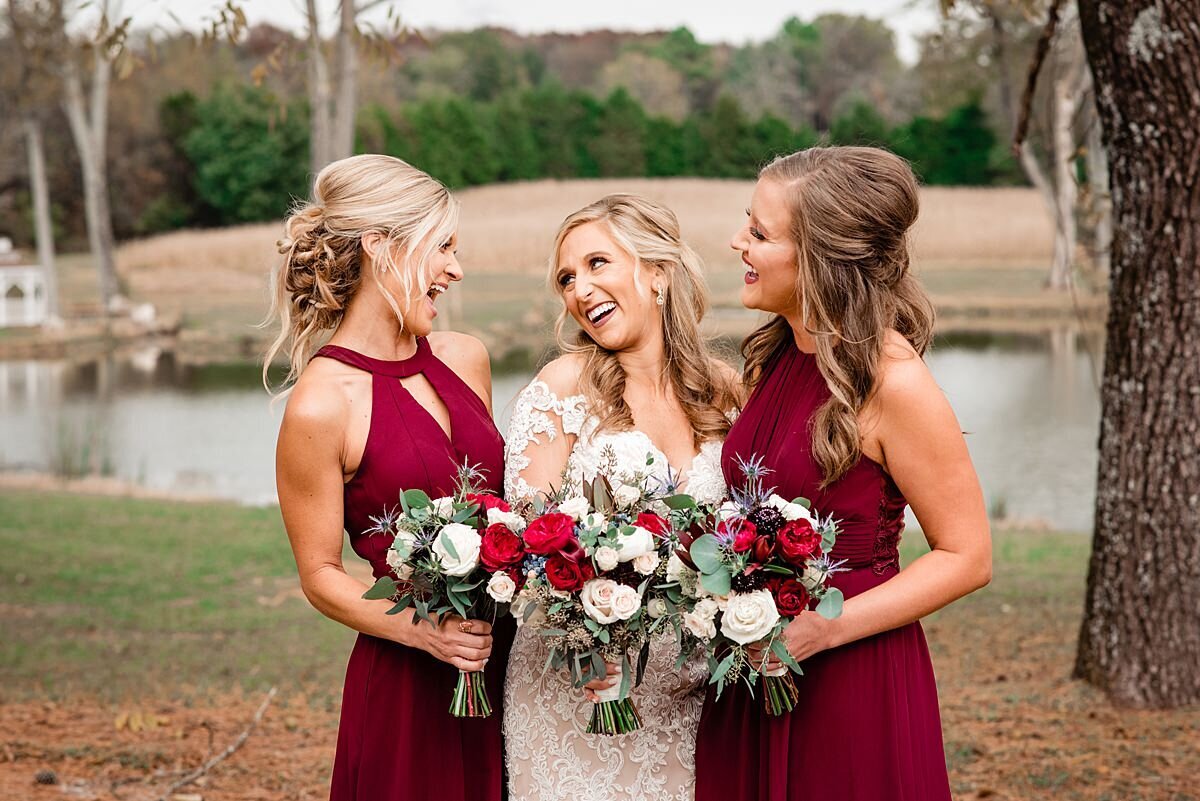 blonde bride wearing a long lace dress laughs with her bridesmaids wearing dark red dresses holding red and white flowers while standing by a lake