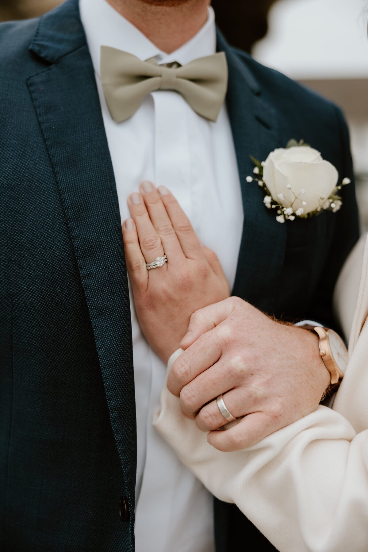 A close-up shot of a bride and groom’s hands showing their wedding rings. The groom is wearing a navy suit with a sage green bow tie and a white rose boutonniere, while the bride’s hand rests on his chest, showing her engagement ring and wedding band. Her arm is wrapped around his, highlighting their connection.