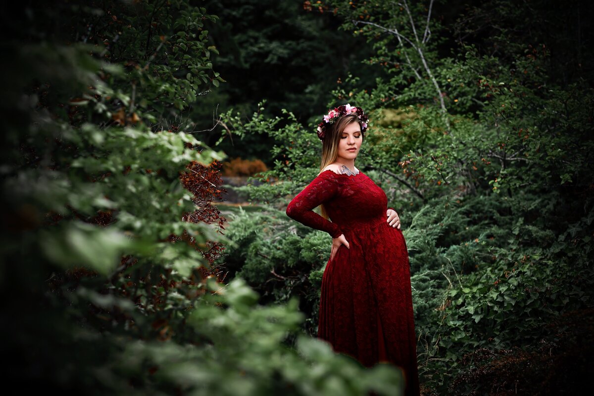 Woman in red dress surrounded by greenery for maternity photoshoot.